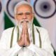 An Indian political leader standing with folded hands in front of national flags during an official event